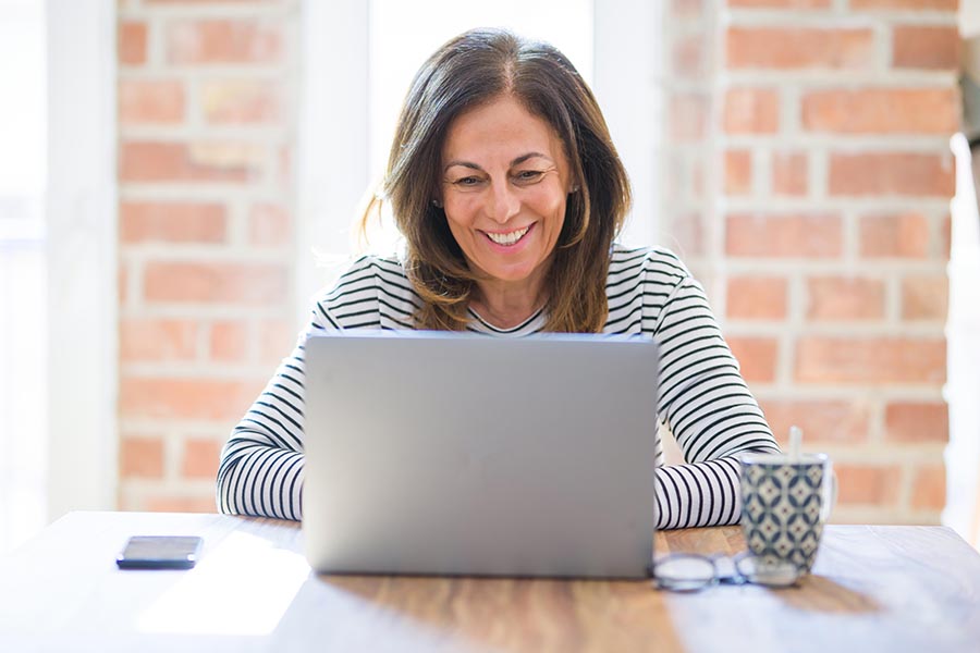Blog - Mature Woman Smiles as She Uses a Laptop at a Wooden Table in Front of a Brick Wall With Coffee and Glasses by Her Side